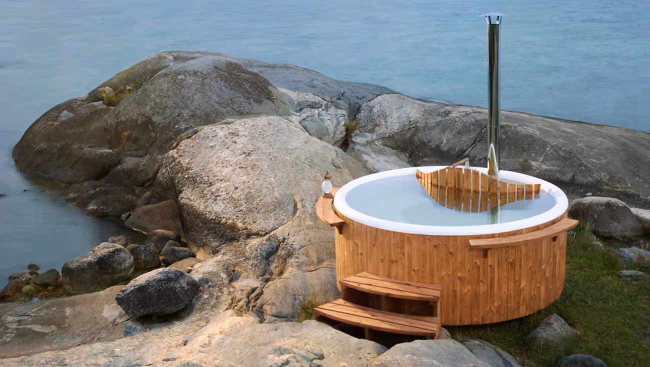 Wooden outdoor hot tub filled with water, placed on rocky terrain near a calm body of water.