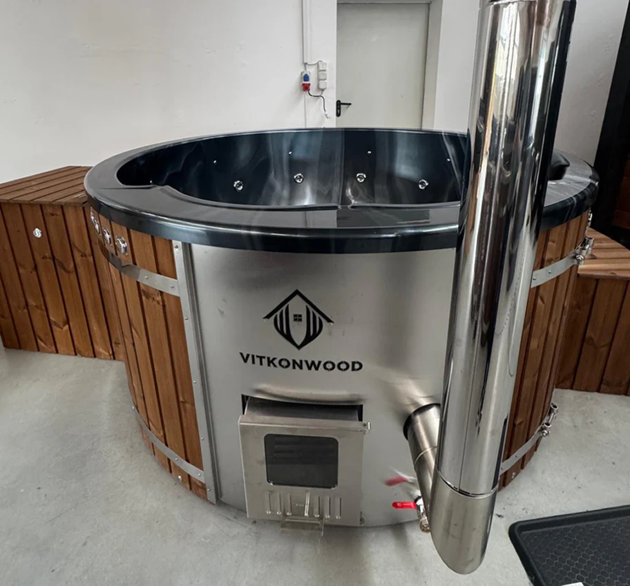 Wooden and stainless steel hot tub with a built-in stove and chimney pipe in a room.