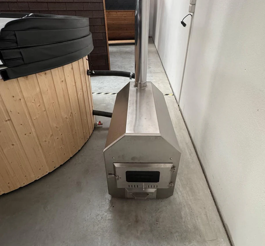 Stainless steel wood-burning stove connected to a wooden hot tub with hoses.