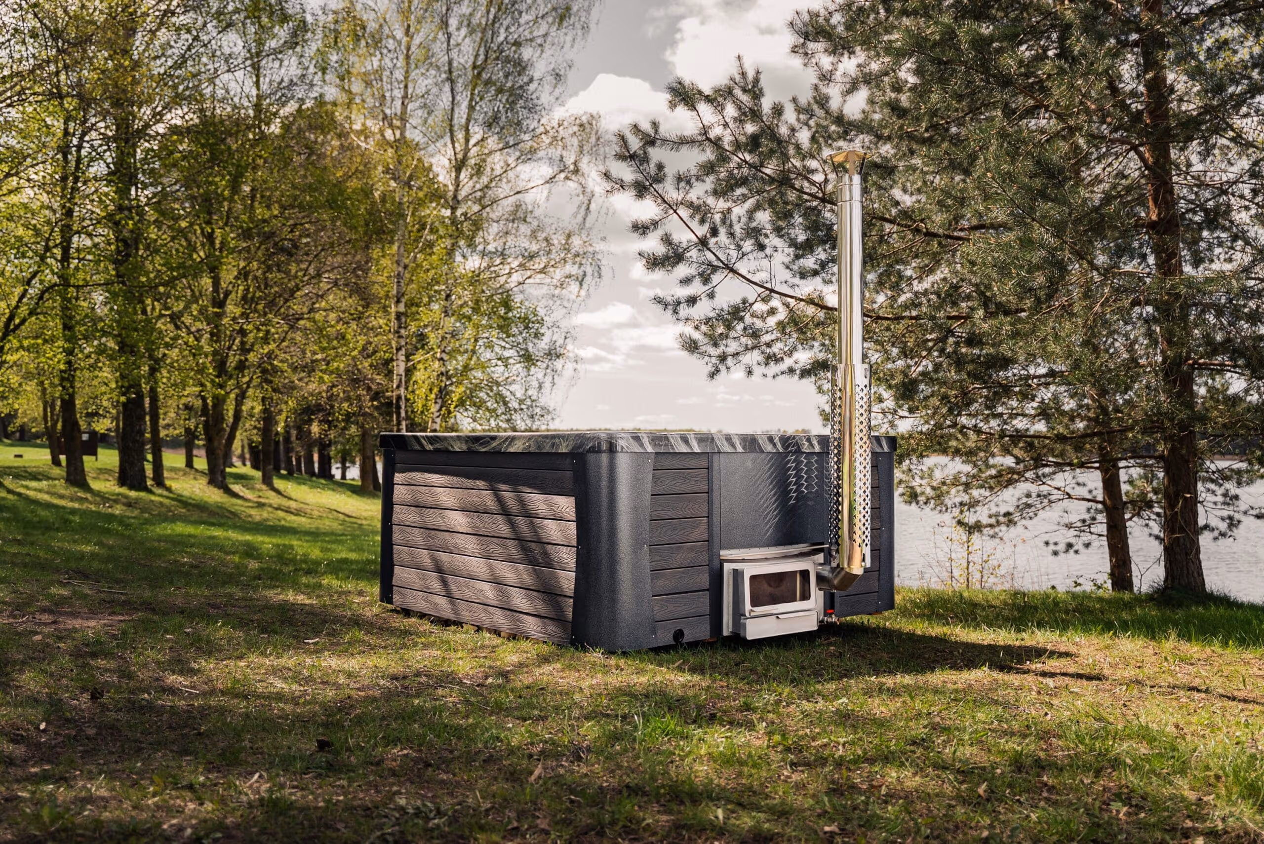 Outdoor wooden hot tub with a metal chimney set on grass beside a lake, surrounded by trees under a partly cloudy sky.