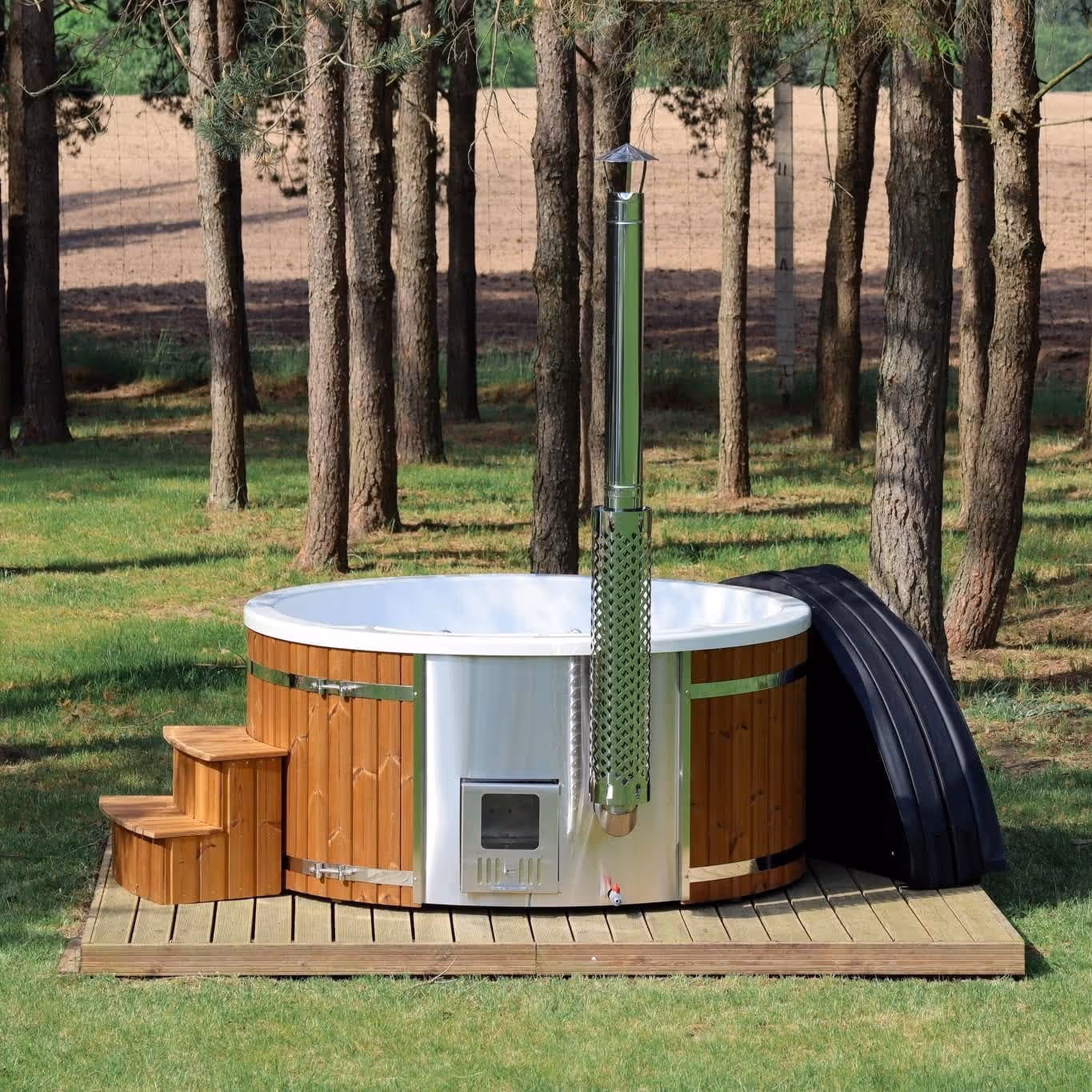Outdoor wooden hot tub with metal firebox and chimney on a wooden deck, surrounded by trees and grass.