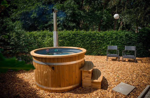 Wooden outdoor hot tub with steps on mulch ground near two black chairs and green hedge background.