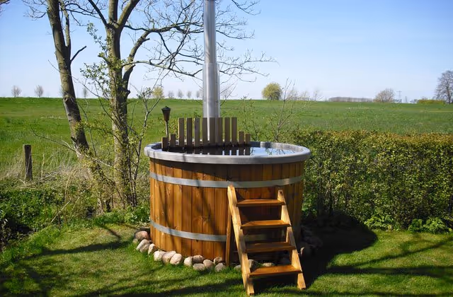 Wooden outdoor hot tub with metal bands, a small wooden ladder, and a chimney, set on grass near trees and a hedge.
