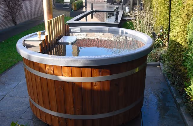 Wooden hot tub filled with clear water, set outdoors on stone tiles surrounded by greenery and a brick wall.