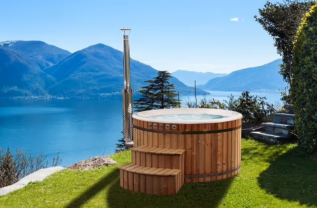 Wooden hot tub with steps on a grassy hill overlooking a lake and mountainous landscape under a clear sky.