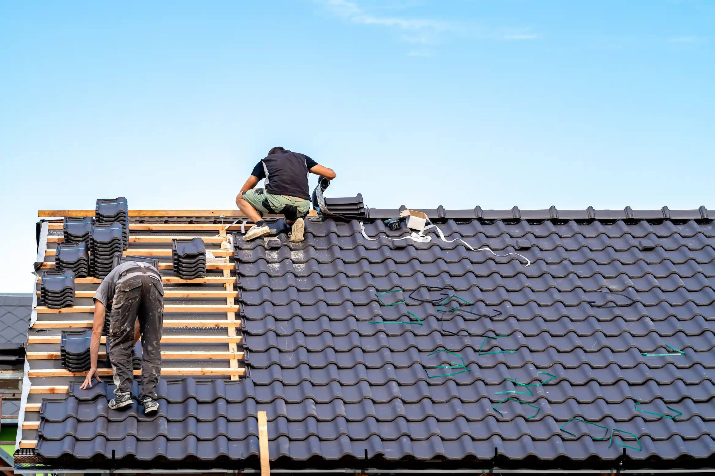 Two workers installing black ceramic roof tiles on a house under clear blue sky.