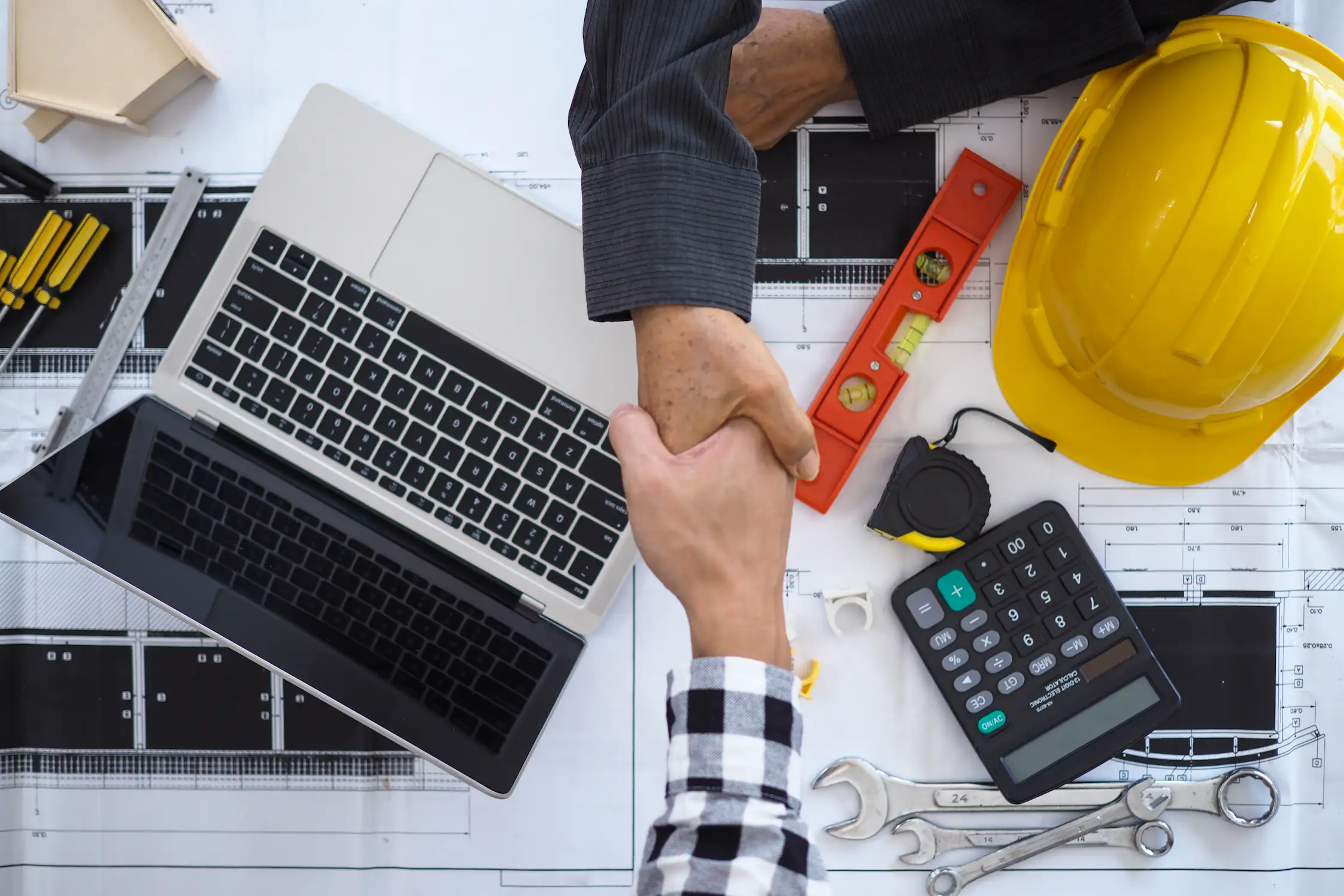 Two people shaking hands over a laptop, blueprints, and construction tools including a yellow hard hat, wrenches, calculator, and level.