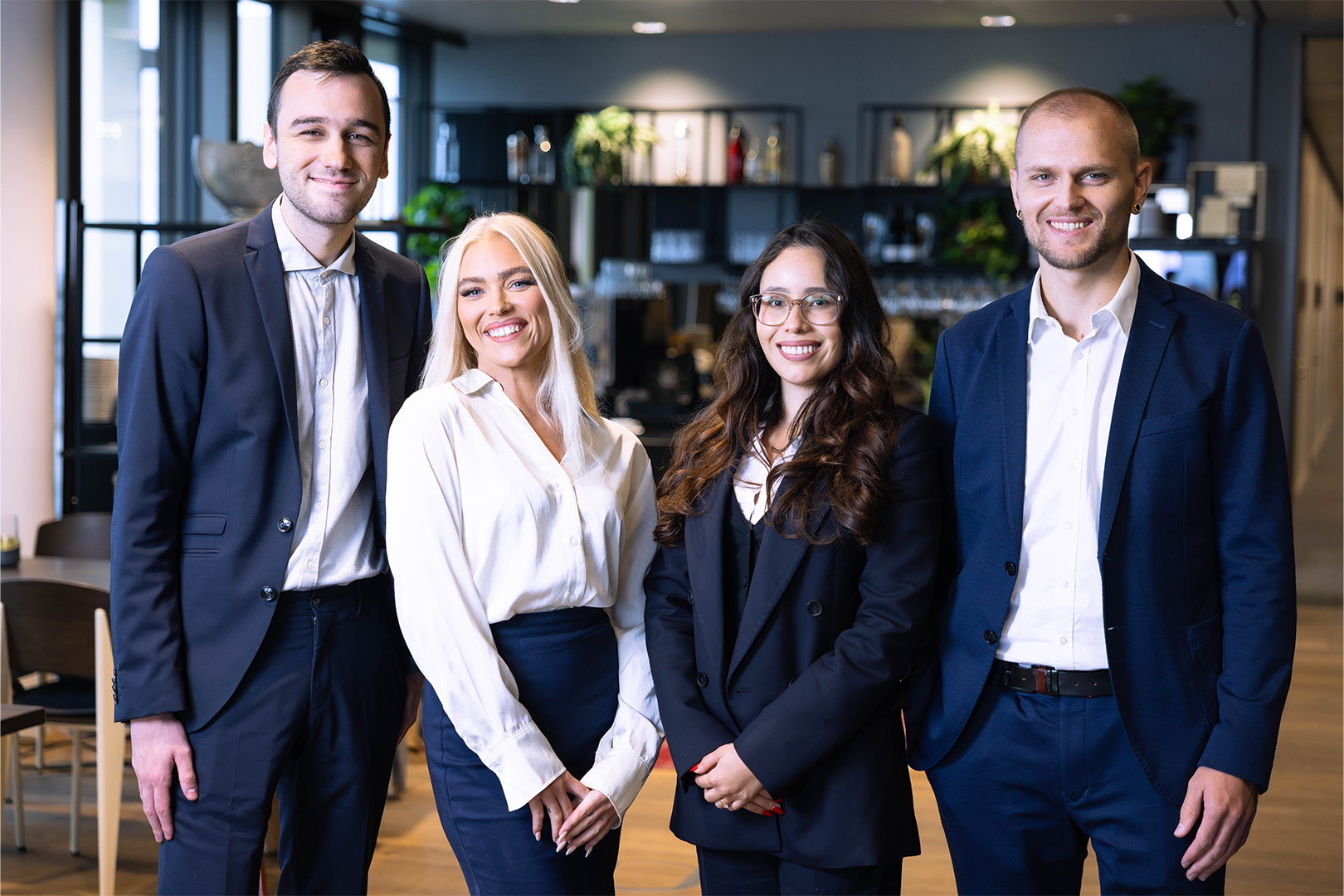 Group of four young professionals smiling, standing indoors in business attire.