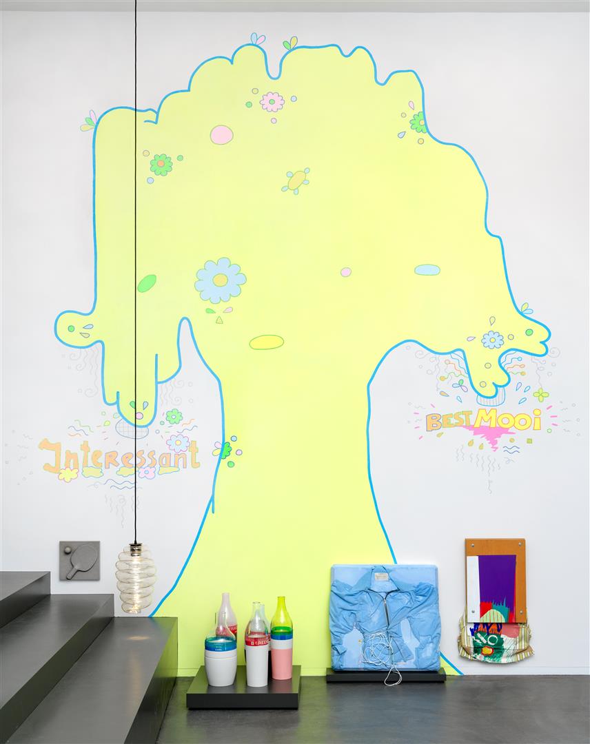 Een muurschildering van een lichtgele boom, met aan de linkerkant de tekst 'interessant' en aan de rechterkant de tekst 'best mooi'. Voor de muurschildering zijn enkele kunstvoorwerpen tentoongesteld. 