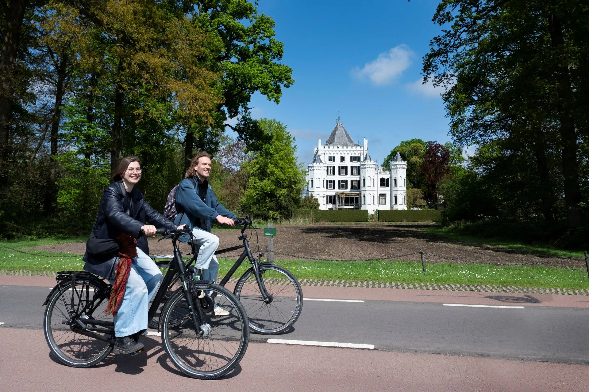 Een vrouw en een man fietsen voorbij aan een wit landhuis