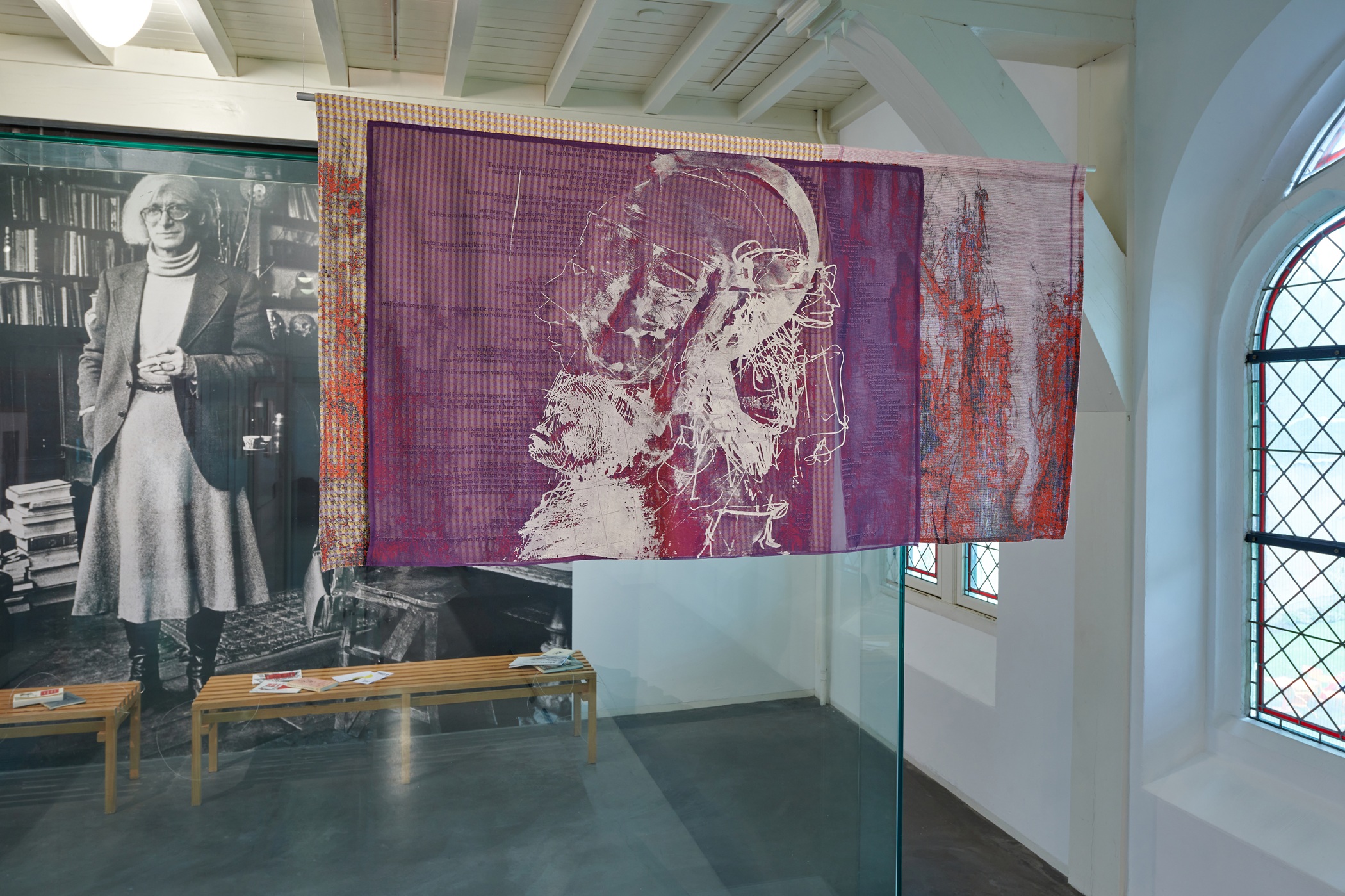 Tentoonstellingszaal, op de voorgrond hangt een wandkleed, op de achtergrond een wandprint van een persoon staand voor een boekenkast