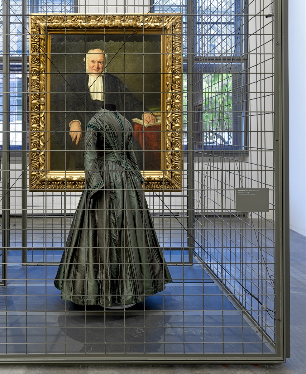 Jurk tentoongesteld in een museumzaal, achter een grijs hekwerk. Achter de jurk hangt een schilderij, een portret van een man.