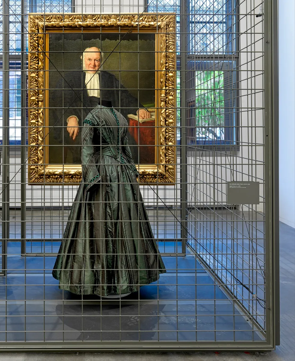 Jurk tentoongesteld in een museumzaal, achter een grijs hekwerk. Achter de jurk hangt een schilderij, een portret van een man.