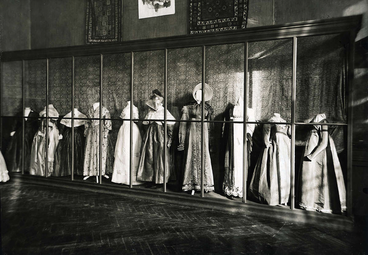 Foto van een verzameling jurken op mannequins, in een grote vitrine met houten latwerk.