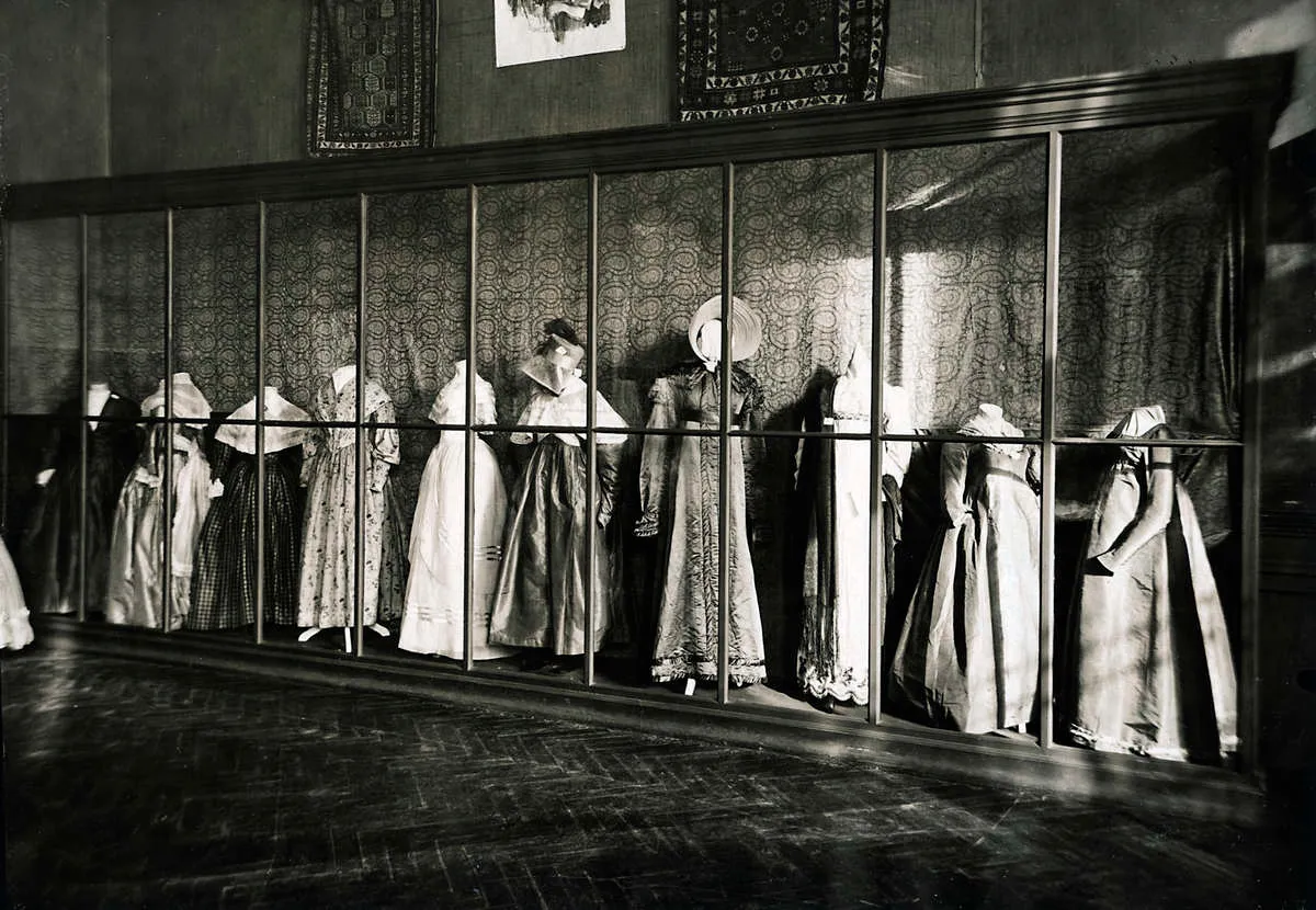 Foto van een verzameling jurken op mannequins, in een grote vitrine met houten latwerk.