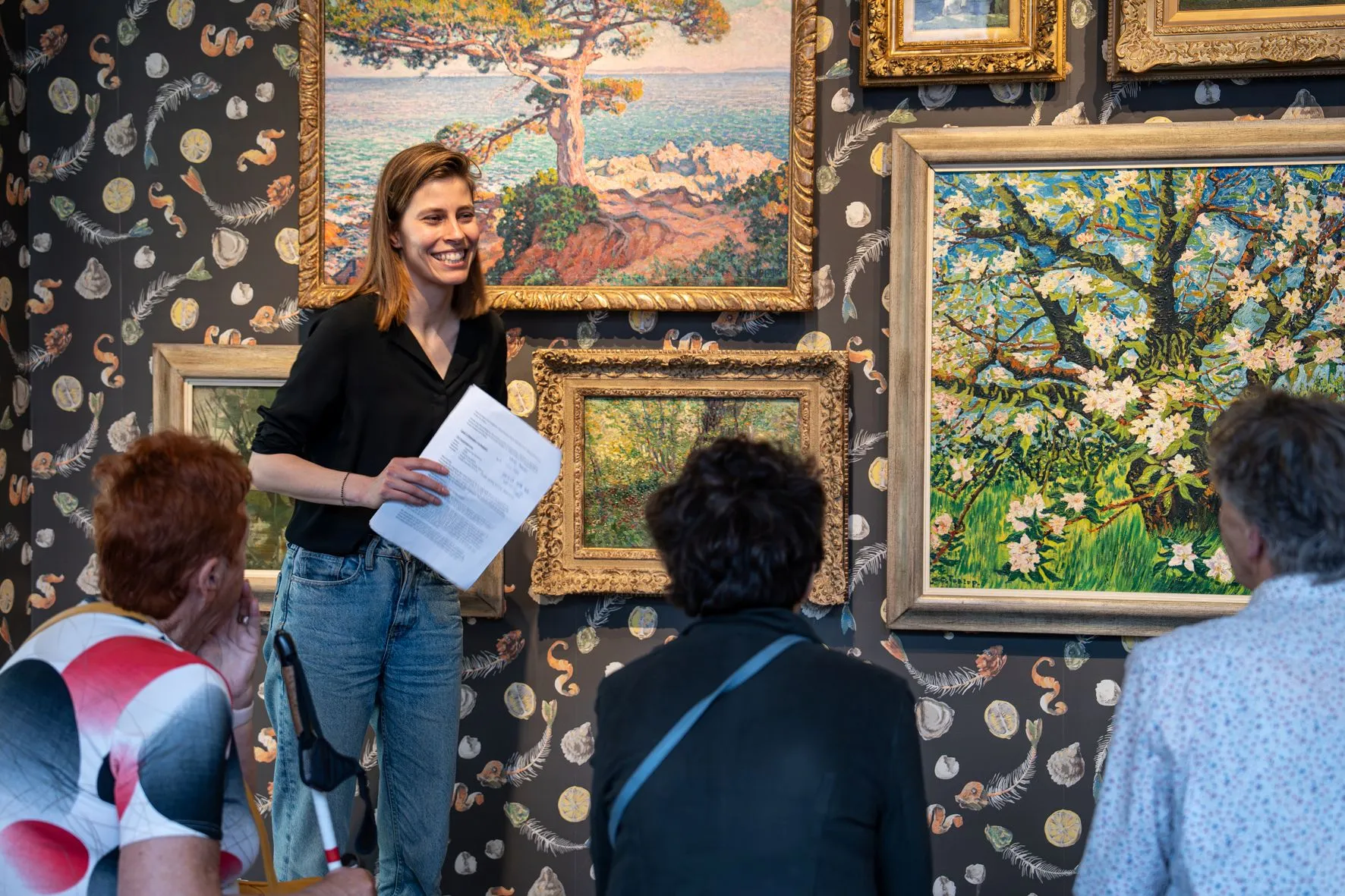 een rondleider staat voor een wand met 4 schilderen. Drie vrouwen luisteren naar haar