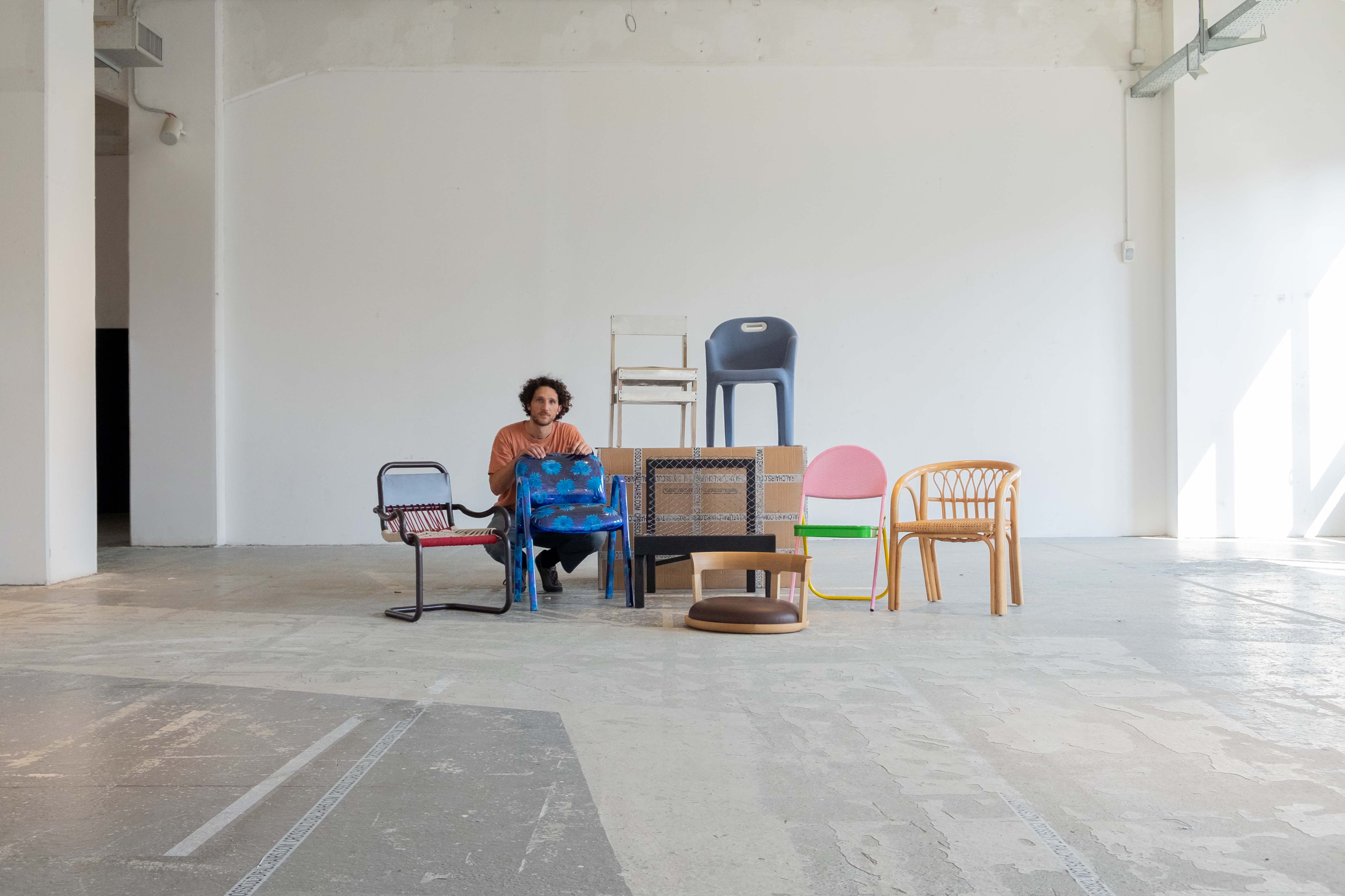 In een grote witte ruimte zit een man gehurkt achter een stoel met blauwe bloemen, Zeven andere, zeer uiteenlopende, stoelen staan naast hem.