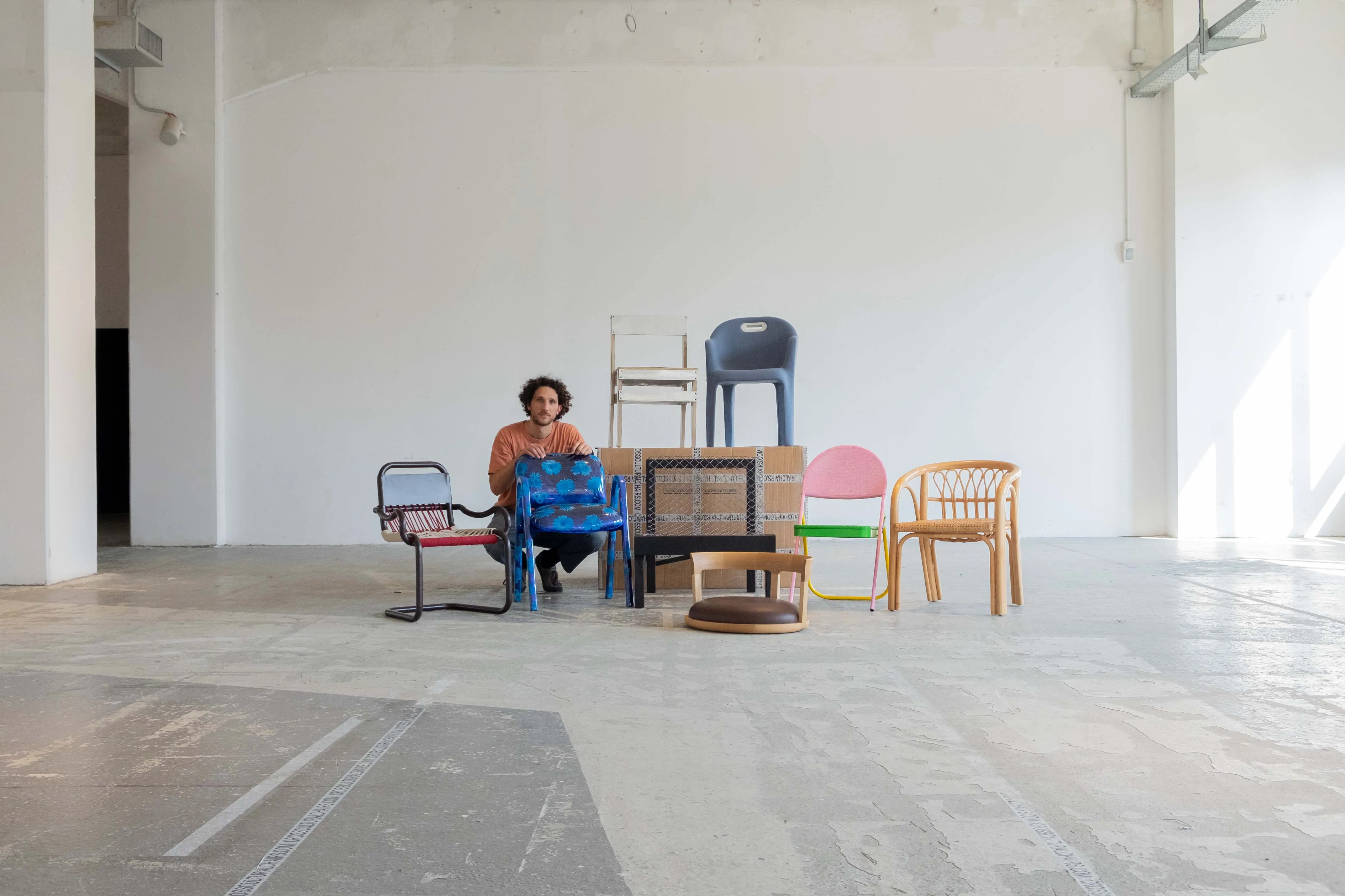 In een grote witte ruimte zit een man gehurkt achter een stoel met blauwe bloemen, Zeven andere, zeer uiteenlopende, stoelen staan naast hem.