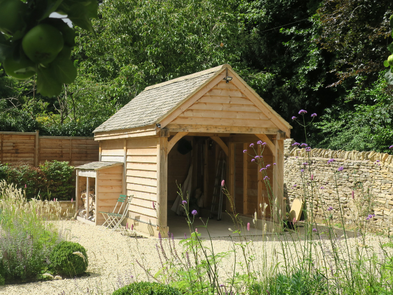 Wooden garden shed with pitched roof in a lush green garden surrounded by flowering plants and a stone wall.