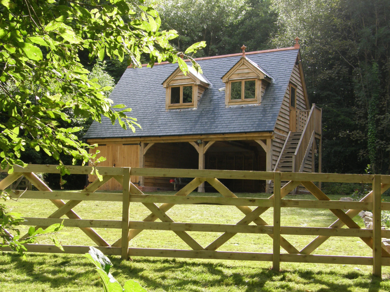A wooden three-bay garage with a room above, featuring two dormer windows and an external staircase, surrounded by greenery and a wooden fence in front.
