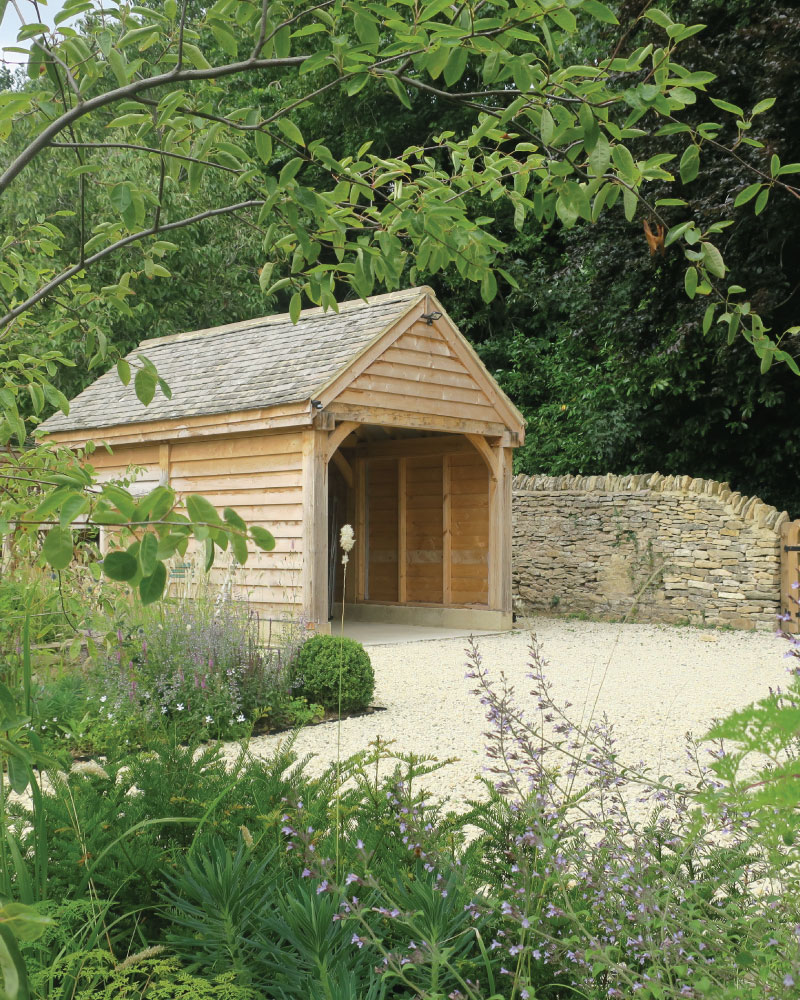 Wooden carport with a gravel driveway, surrounded by green plants and a stone wall in the background.