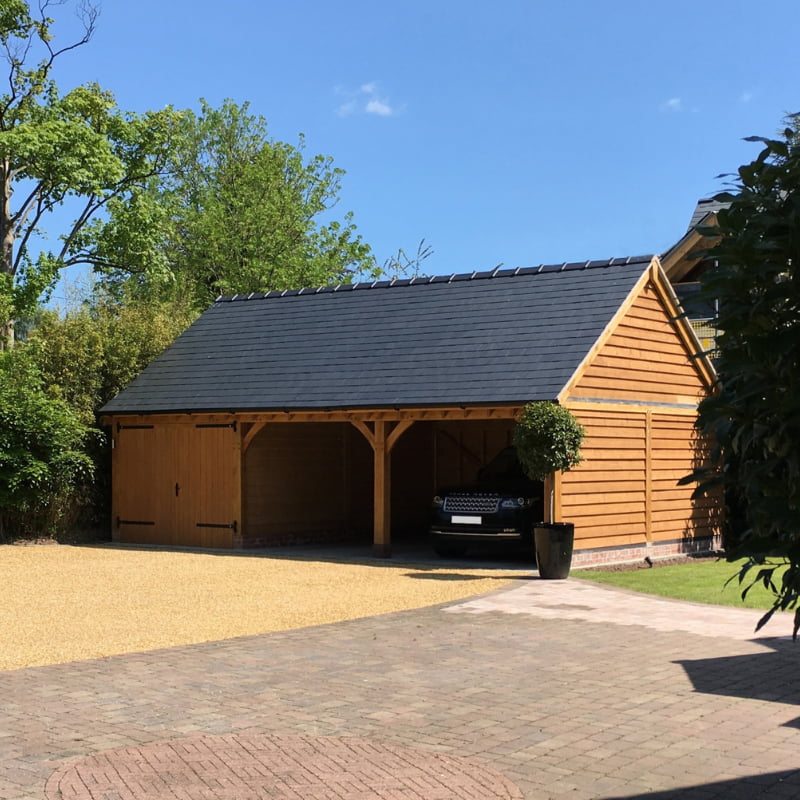 Single-storey oak-framed garage with a slate roof, surrounded by trees and brick driveway