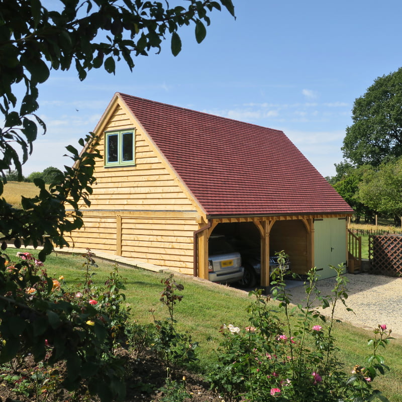 Wooden garage with oak frame, red tiled roof, and a small green window, surrounded by a garden with flowers under a clear blue sky.