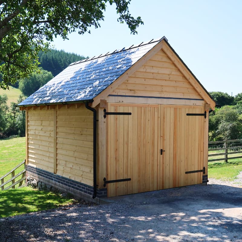 Single-storey oak-framed garage with a slate roof, surrounded by trees and has garage doors with ironmongery fittings.