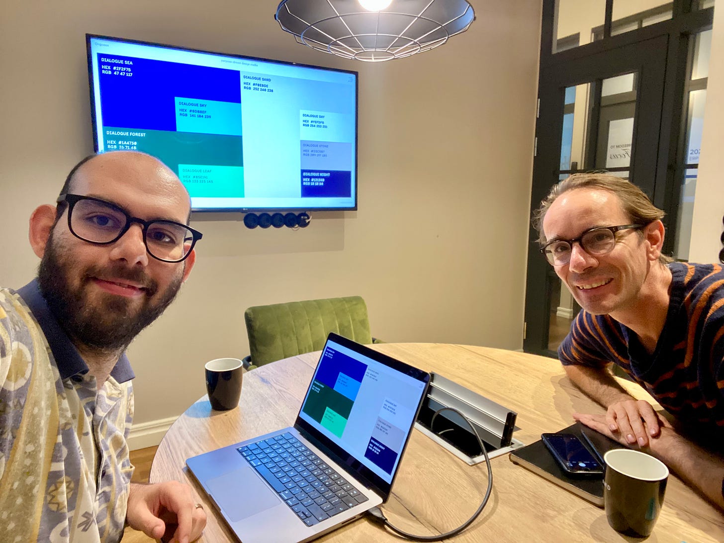 Savvas smiling in an office with a round table with Estonian's Dialogue CEOa A laptop and a large wall screen display multicolored charts. The mood is collaborative and friendly.