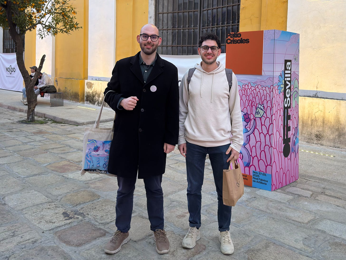 Antonis Theodosiou and Savvas Theodosioui at OFFSevilla Festival. stand smiling on a cobblestone street. One wears a black coat, the other a beige hoodie. Behind them is a large colorful mural with the word "OFFSevilla." The setting is bright and cheerful.