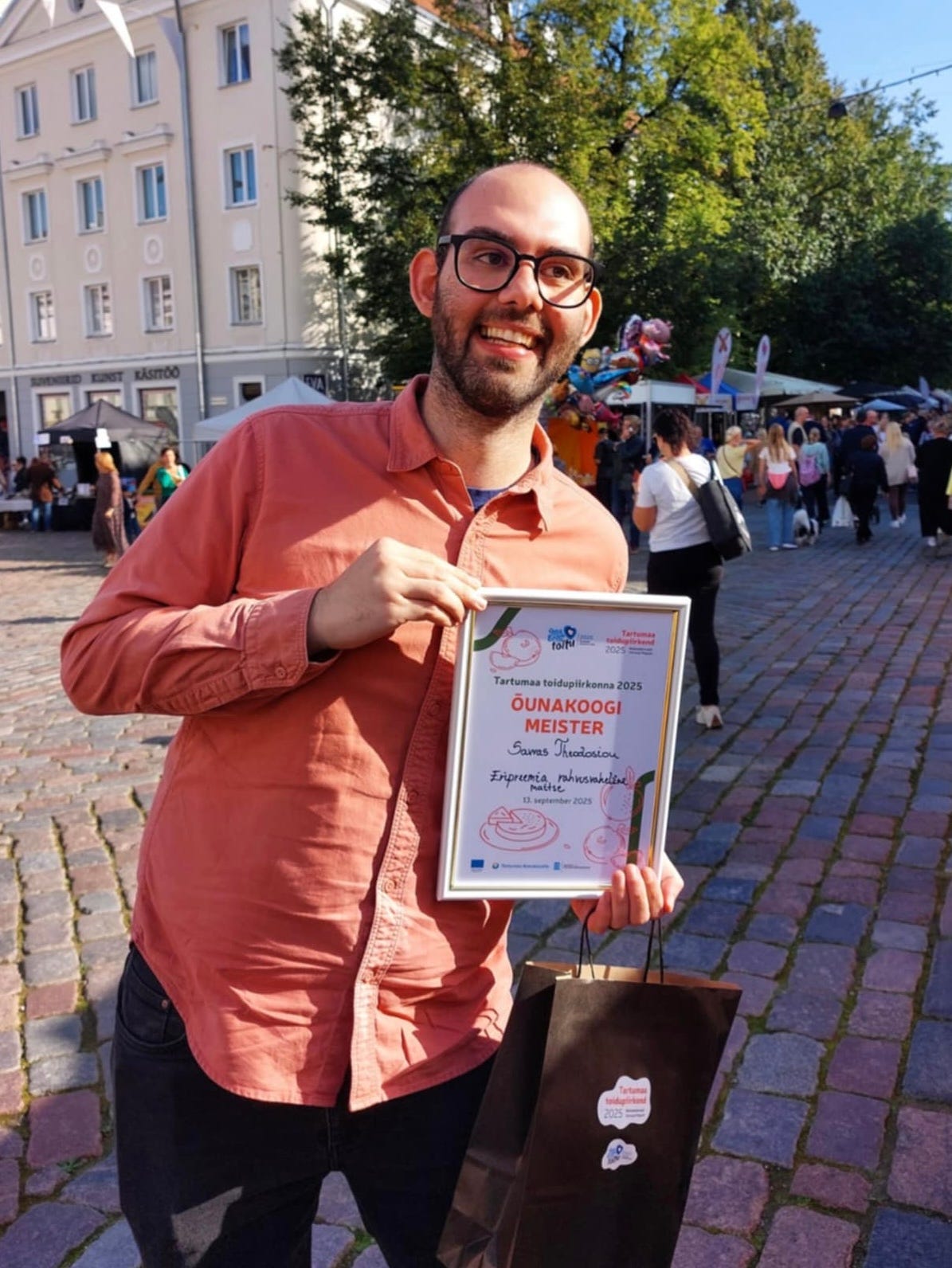 Savvas in a coral shirt smiles and holds a framed certificate for a special award for international taste at the local apple pie competition in Estonia. He is outdoors in a sunlit cobblestone plaza. People and a building are visible in the background.