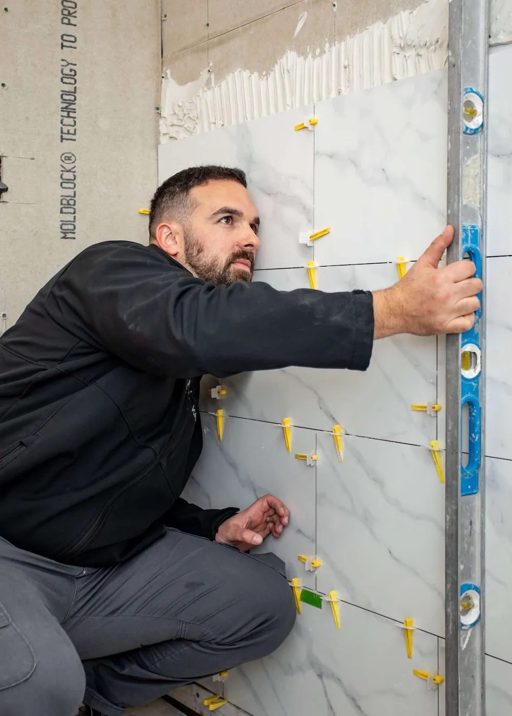 Contractor tiling a bathroom wall with marble-style tiles and precision leveling tools