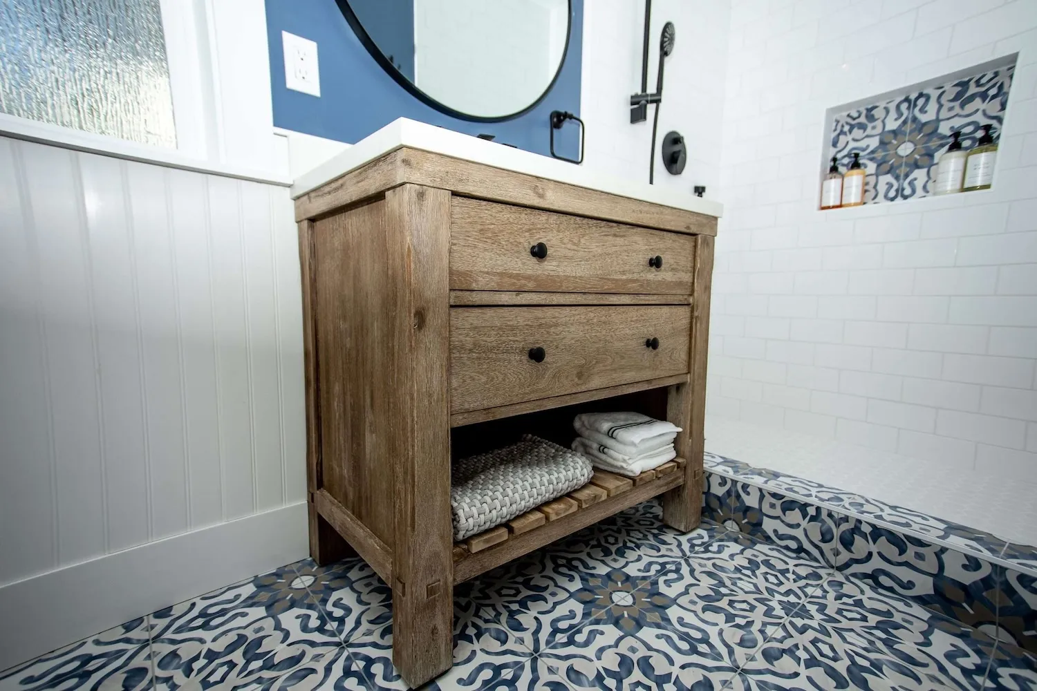 Rustic wood vanity with towels and black hardware in a bathroom with decorative tile floor and white tile shower