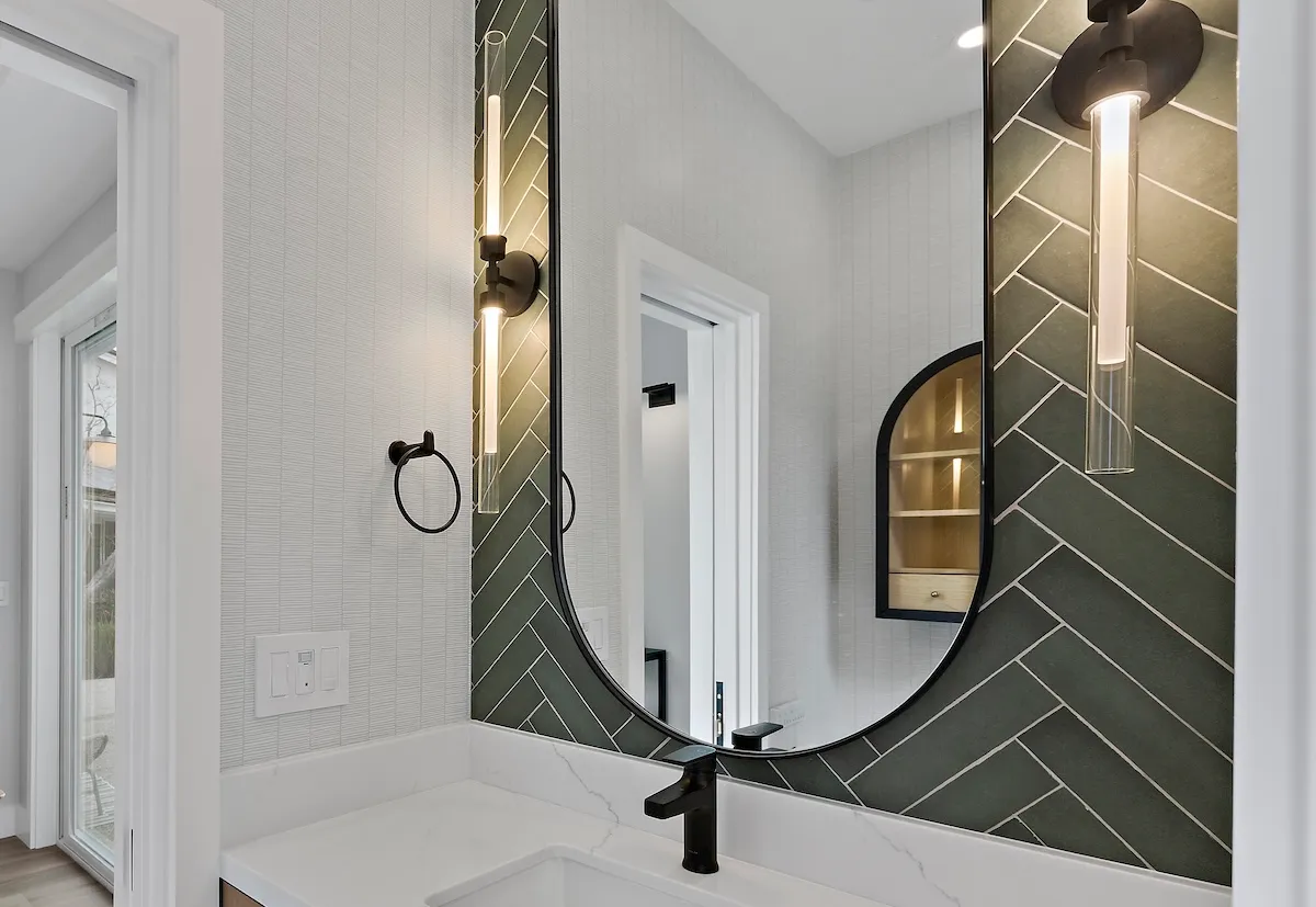 Modern bathroom vanity with a green herringbone tile accent wall, circular mirror, and vertical wall lights