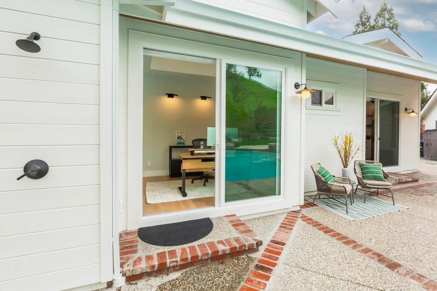 White home exterior with sliding glass doors leading to a patio area, showing outdoor seating and a view inside an office space