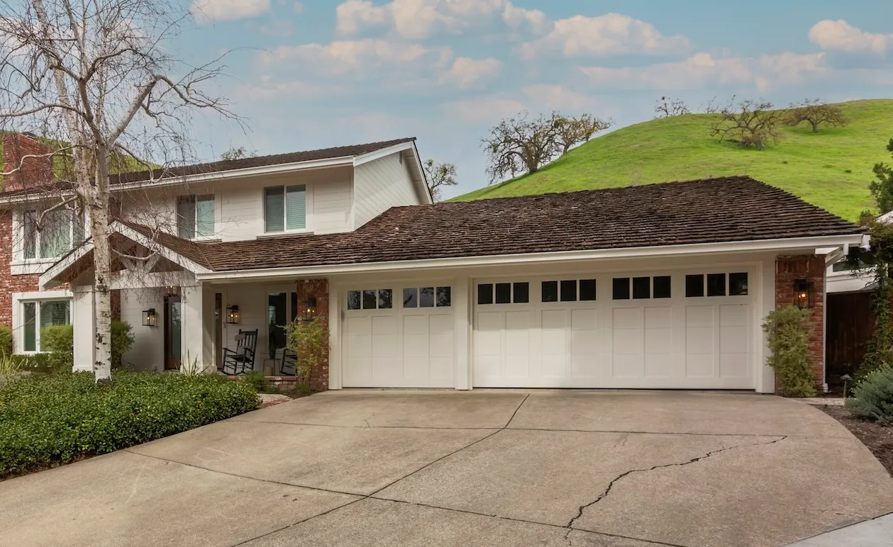 Exterior view of a two-story house with a triple garage and pitched roof, set against a grassy hill backdrop