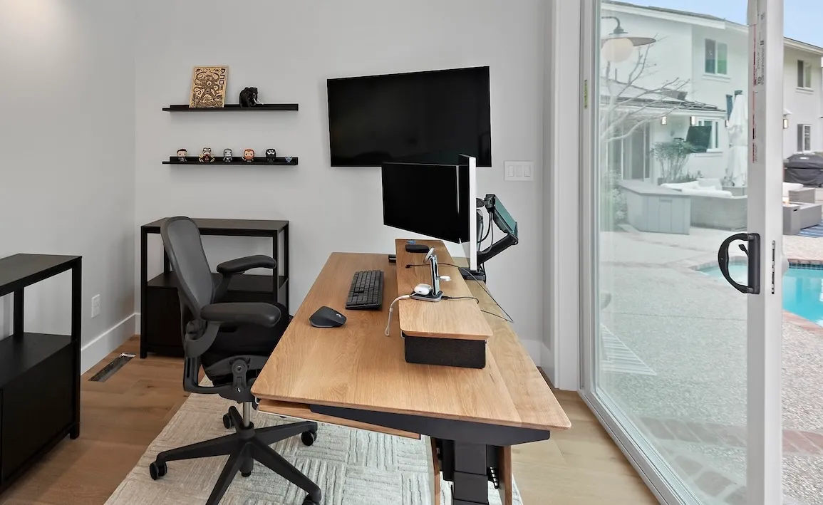Home office setup with a wooden desk, dual monitors, black office chair, and a view of a pool through the sliding door