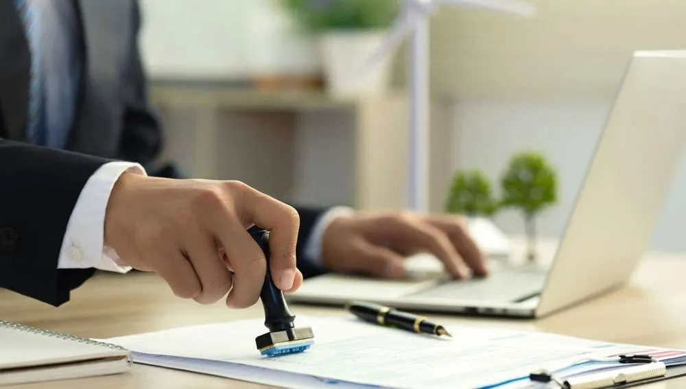 Person in formal attire stamping paperwork at a desk with a laptop, representing contract approval or signing.
