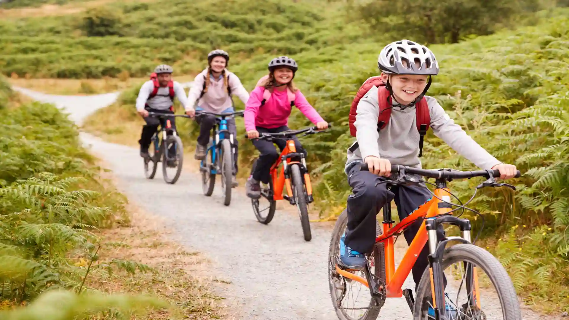 Family group of children and adults riding mountain bikes along a forest trail, smiling and wearing helmets on a gravel path surrounded by greenery.