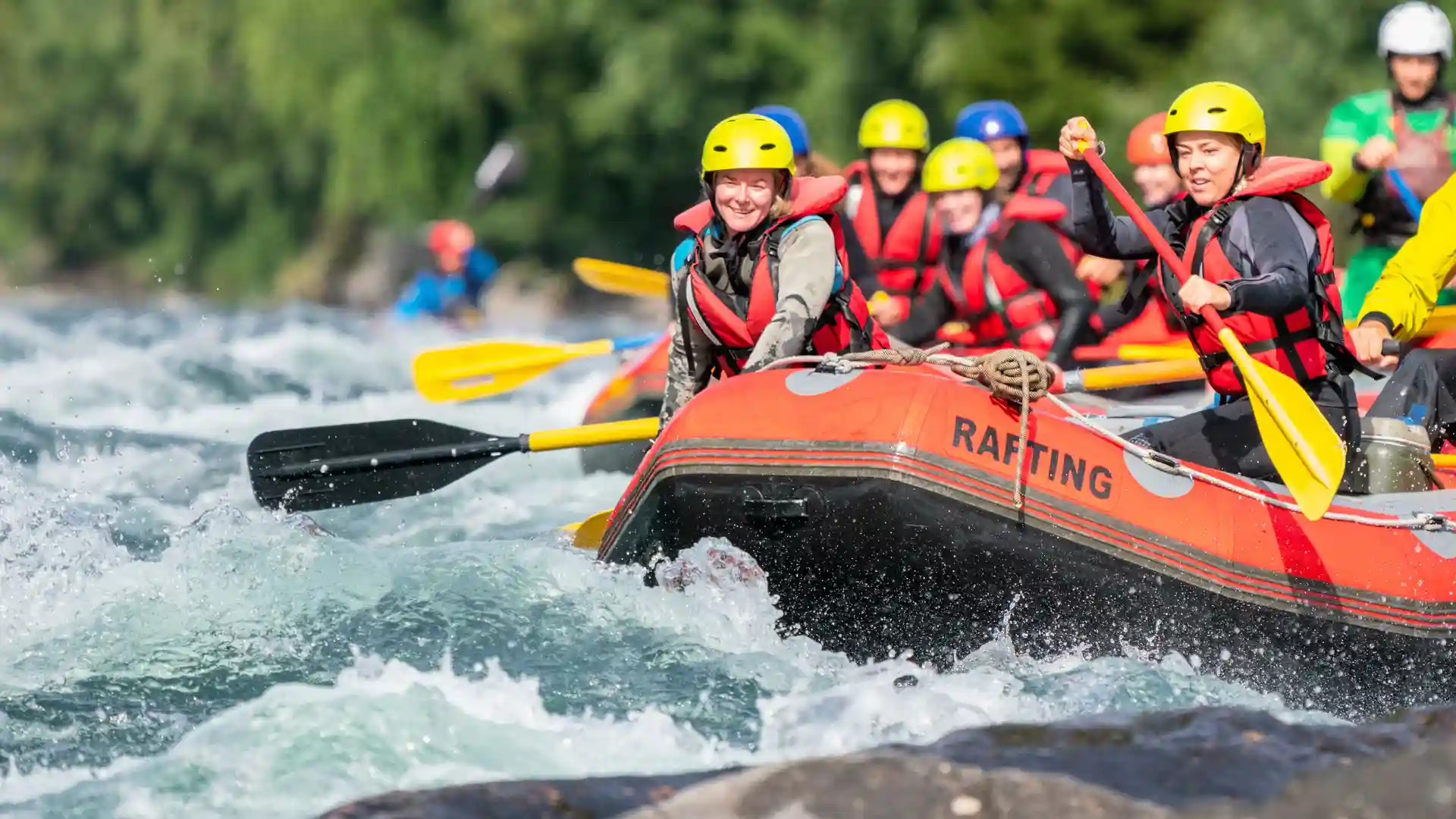 Group of people in helmets and life jackets white-water rafting down a fast-flowing river, navigating rapids in an inflatable raft with paddles.
