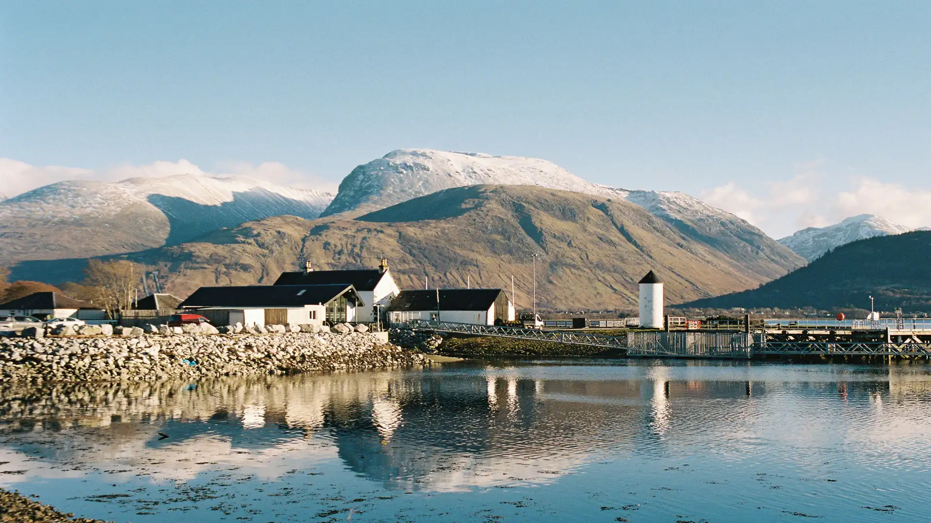 View across Lochaber towards Ben Nevis and Fort William, Scotland’s Outdoor Capital, with reflections on calm water under clear skies.