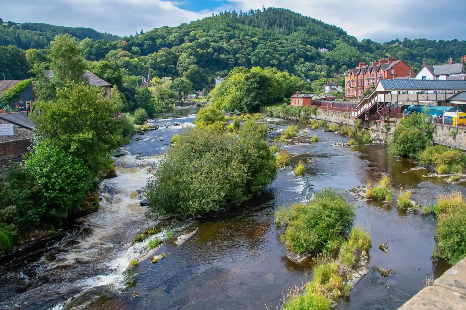 River Dee flowing through Llangollen, North Wales, with heritage railway station, red-brick buildings, and forested hills in the background.