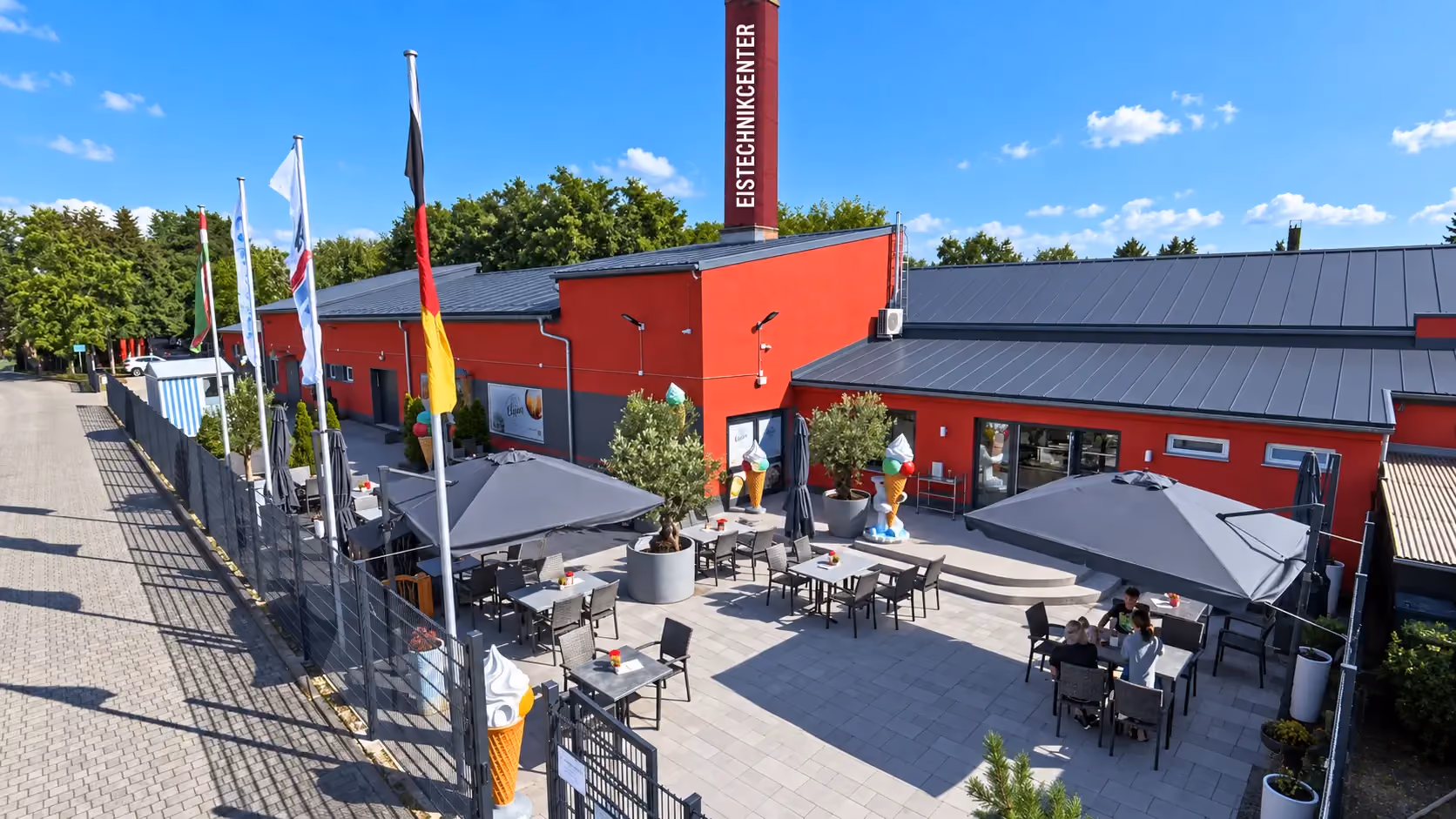 Visualisierung des Eistechnikcenter-Außenbereichs in Luckenwalde mit anthrazitfarbenem Dach, gepflasterter Terrasse und Werbung am Schornstein