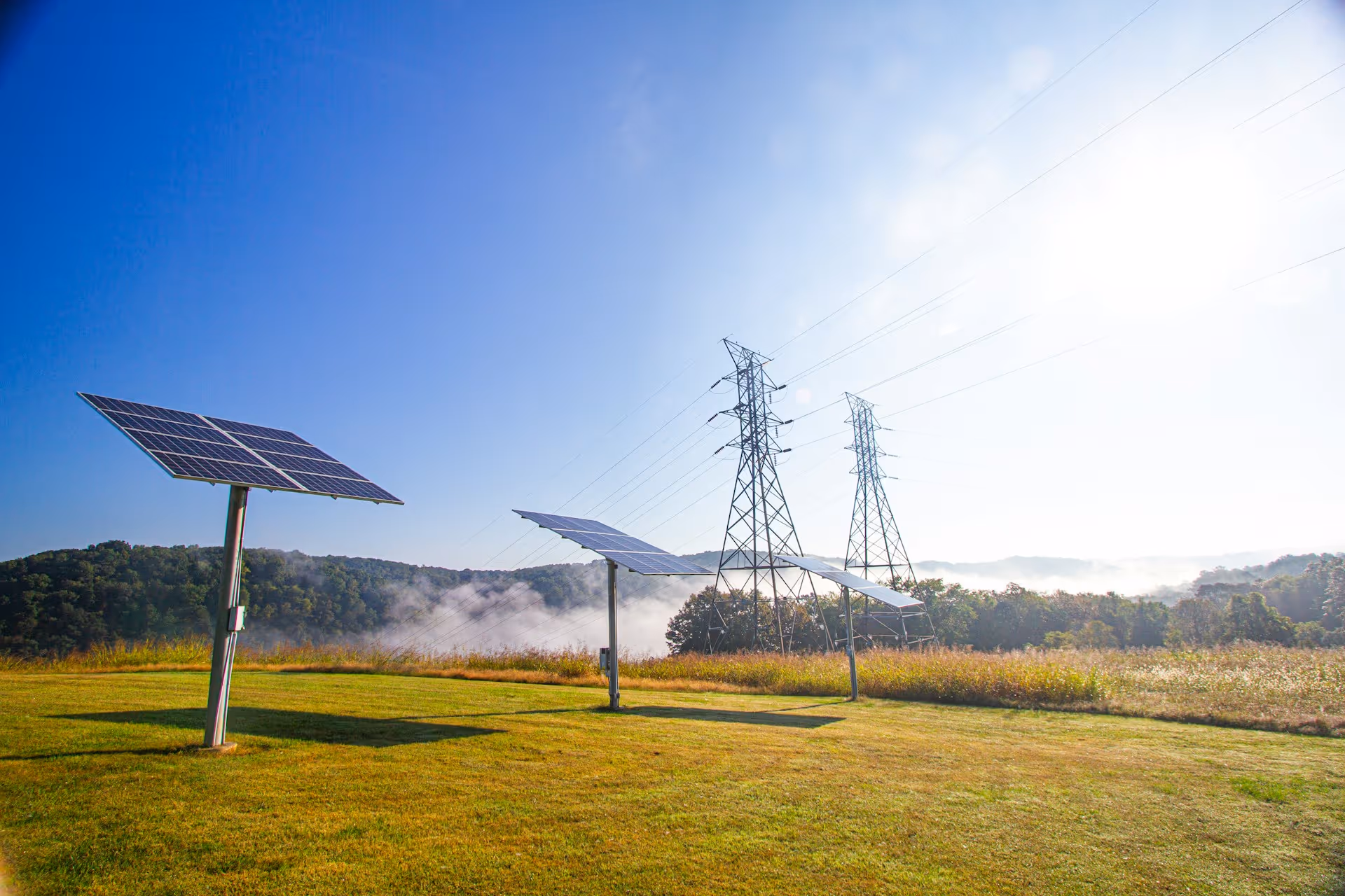 Ground-mounted solar tracking panels stand in a sunlit green field
with morning mist and high-voltage transmission towers in the distance,
representing LONGi's high-efficiency solar technology feeding clean
energy directly into the electrical grid.