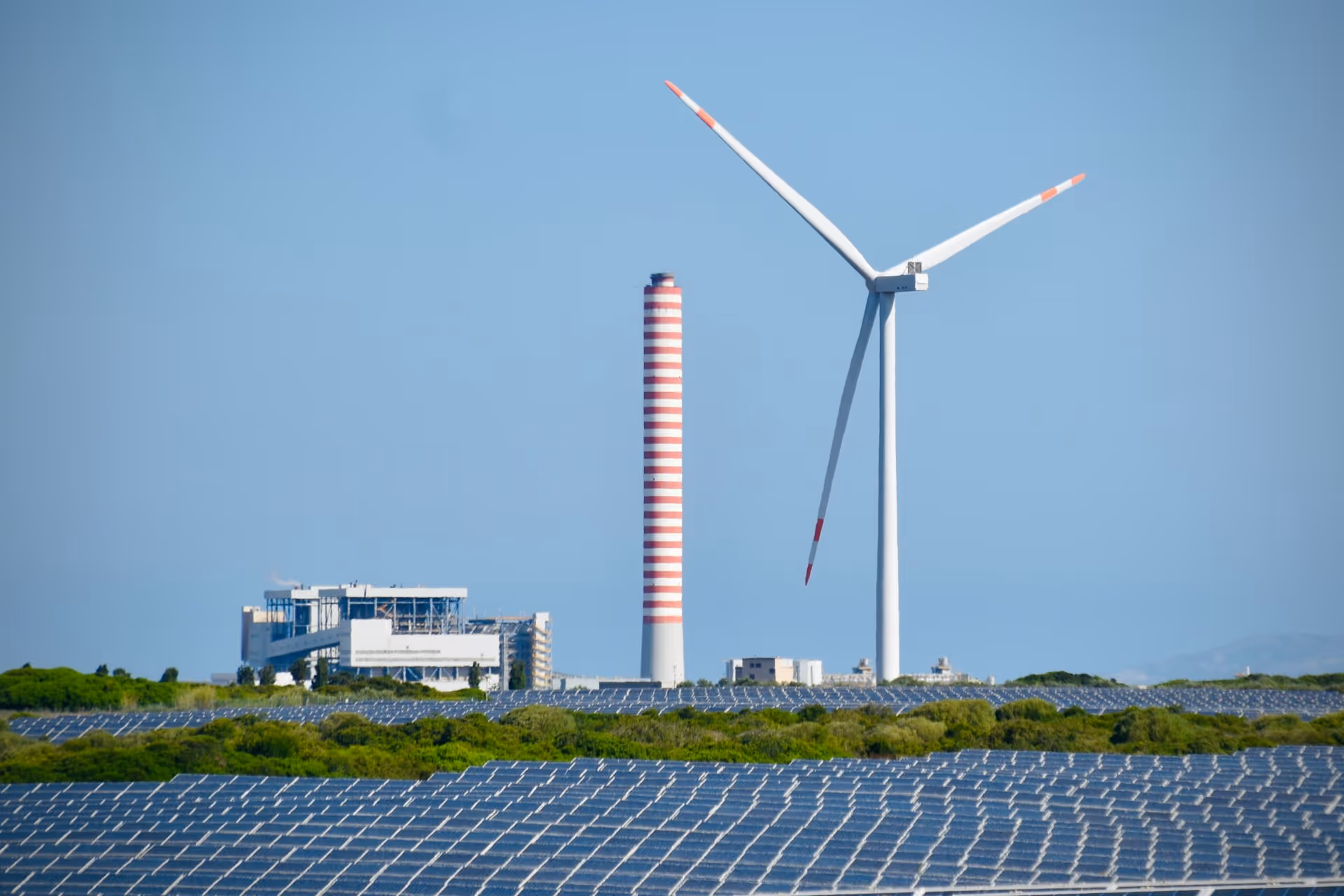 A utility-scale solar farm stretches across the foreground while a
wind turbine and industrial chimney rise in the background under a
clear blue sky — illustrating the energy transition at the heart of
LONGi's climate and societal impact mission.