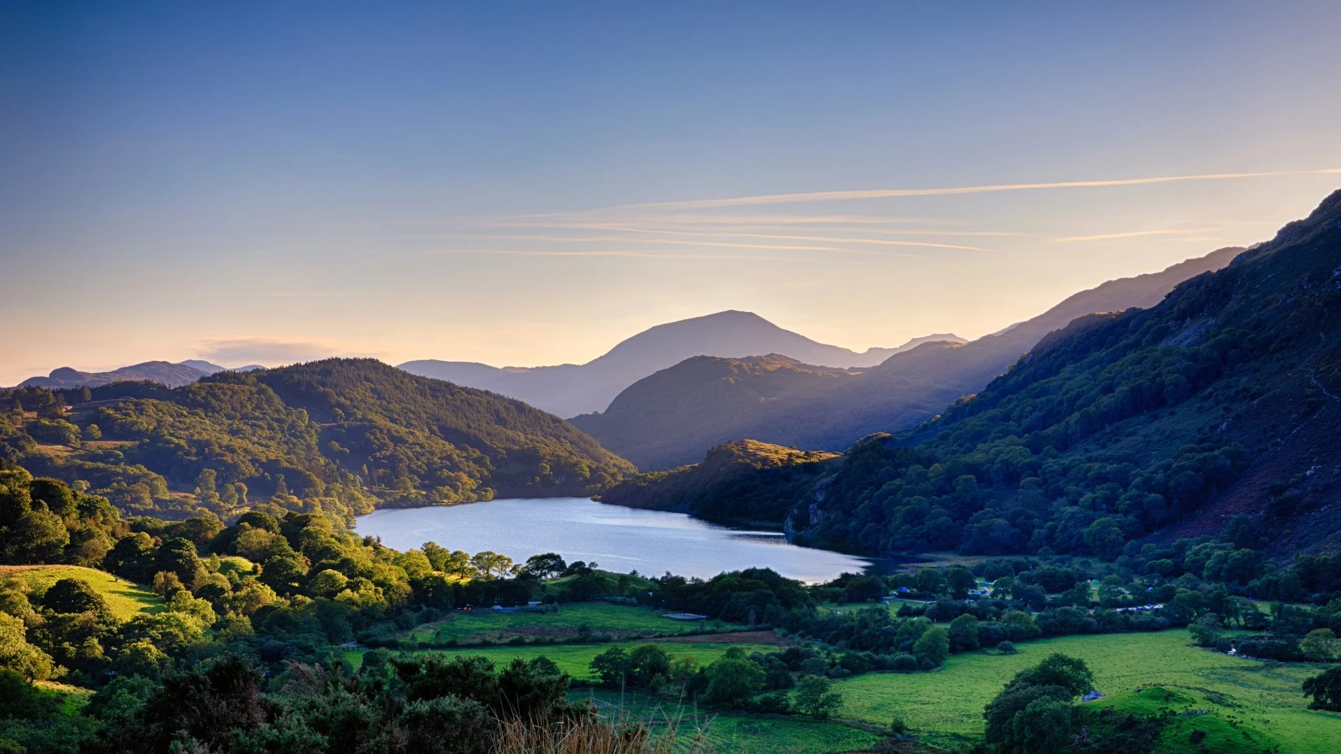 Dramatic landscape of Snowdonia National Park in Wales at sunset, with a winding lake nestled among rolling green hills and rugged mountains under a clear twilight sky.