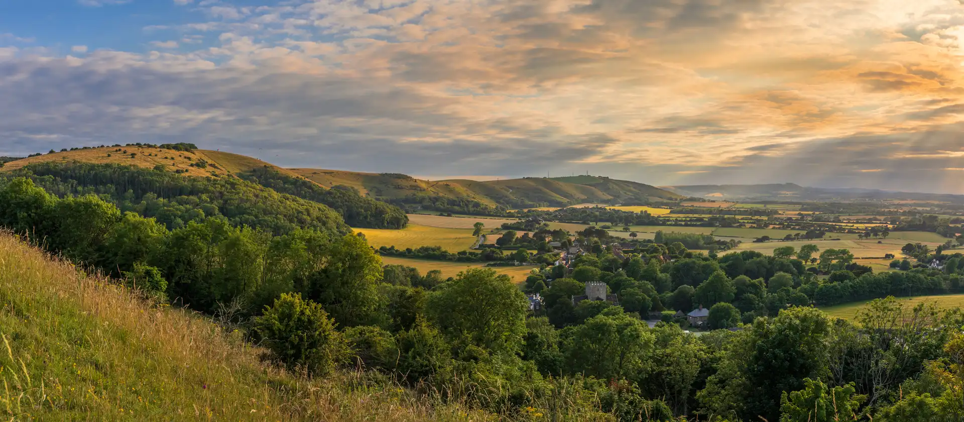 Sunset over a lush English countryside valley, with rolling green hills, patchwork fields, and a small village nestled among trees.