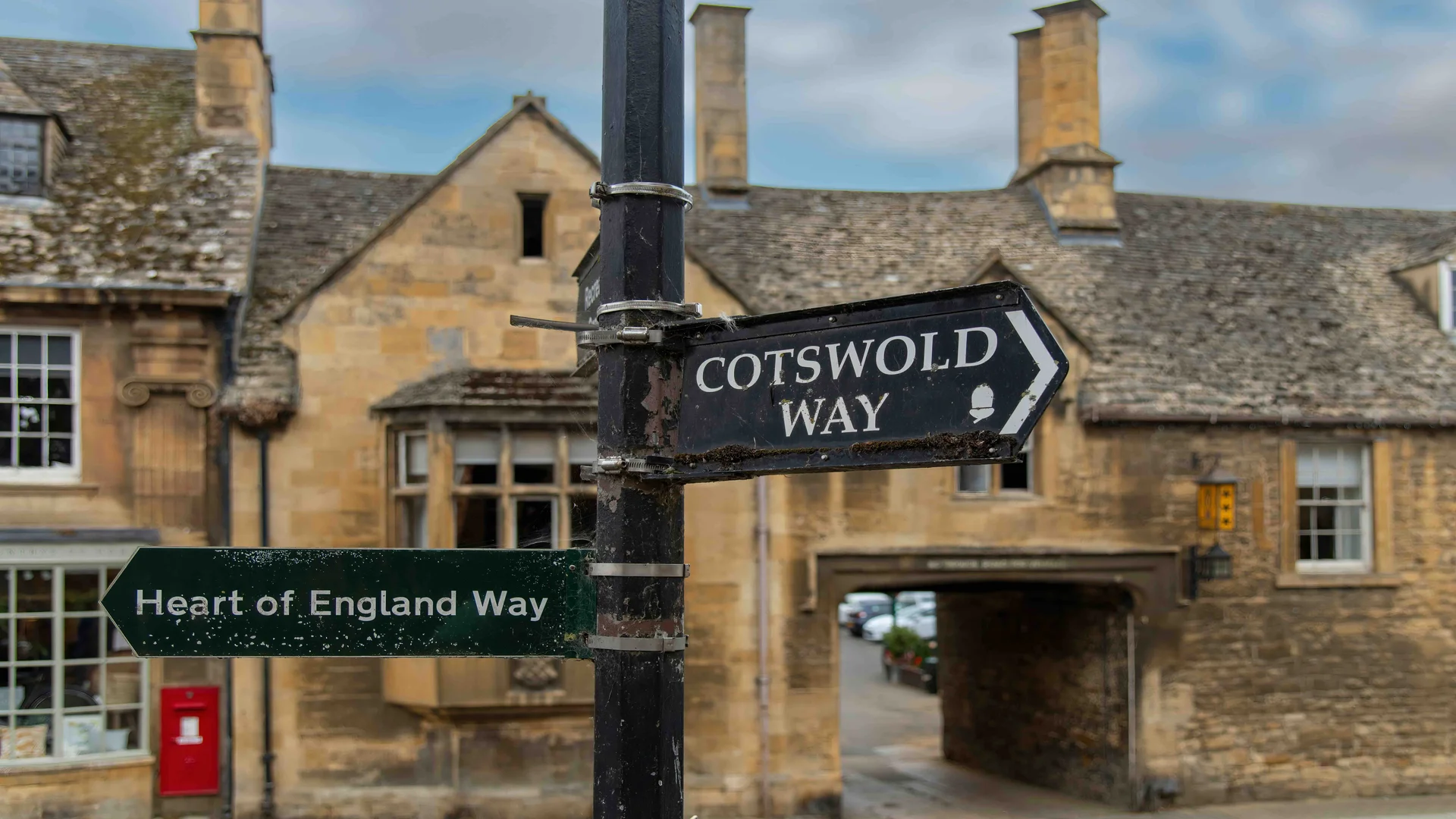 An old sign showing the Cotswolds way stands in front of golden stone buildings.
