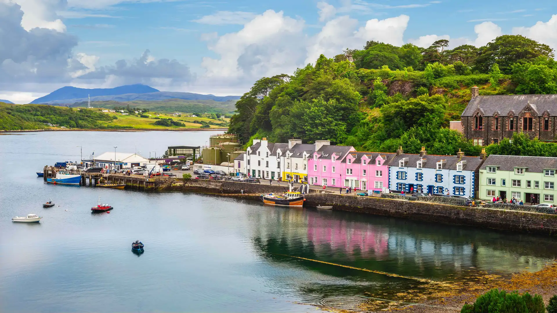 View of Portree Harbour on the Isle of Skye, Scotland, with colourful waterfront houses, moored boats, and green hills under a bright sky.