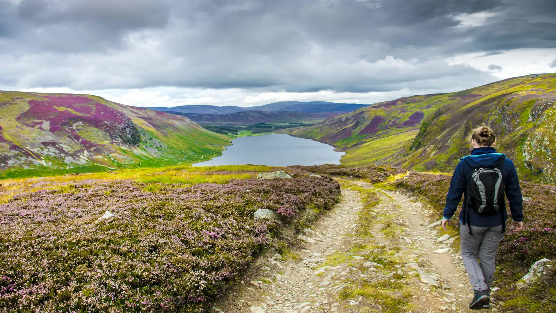 Walker on a heather-lined upland trail in North East Scotland, overlooking a remote loch and surrounding hills under a moody sky.